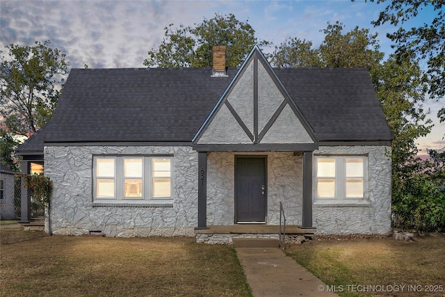 tudor house with roof with shingles, stucco siding, a chimney, a front lawn, and stone siding