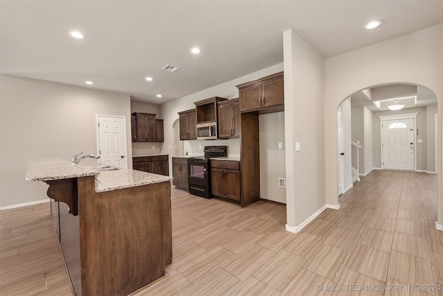 kitchen with an island with sink, light stone counters, sink, dark brown cabinets, and black range with electric stovetop