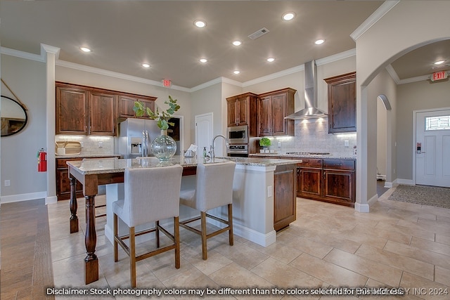 kitchen with light stone counters, a kitchen island with sink, wall chimney range hood, backsplash, and crown molding