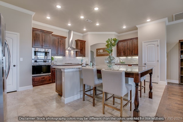 kitchen featuring an island with sink, backsplash, wall chimney exhaust hood, light stone countertops, and stainless steel appliances