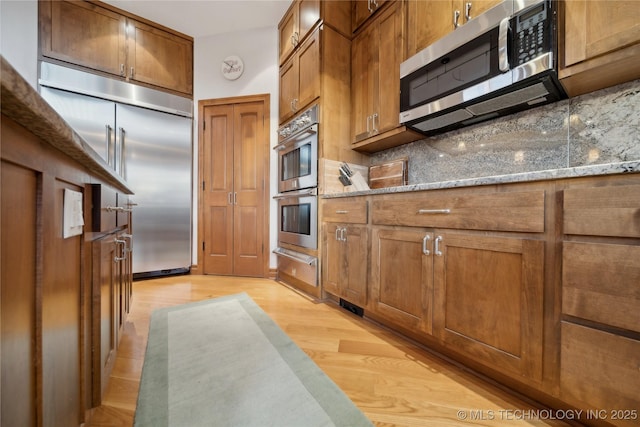 kitchen with stone countertops, appliances with stainless steel finishes, backsplash, brown cabinets, and a warming drawer