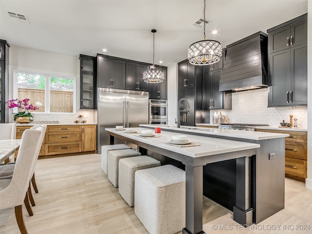 kitchen with decorative light fixtures, custom exhaust hood, stainless steel appliances, and an island with sink