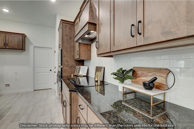 kitchen featuring tasteful backsplash, dark stone counters, stainless steel oven, premium range hood, and black electric stovetop