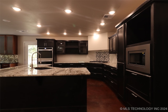 kitchen with decorative backsplash, a kitchen island with sink, stainless steel appliances, sink, and light stone countertops