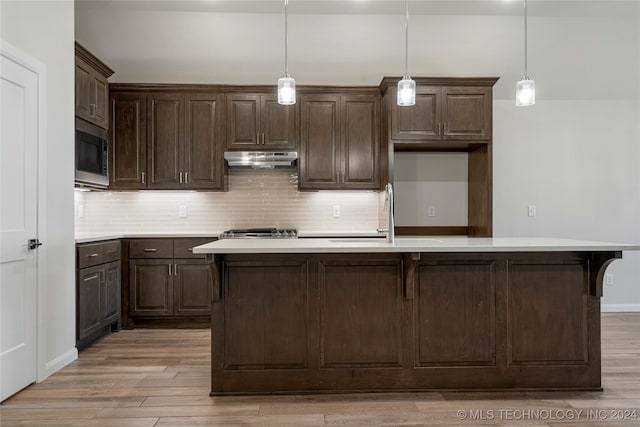 kitchen with sink, a kitchen island with sink, decorative light fixtures, stainless steel microwave, and light hardwood / wood-style floors