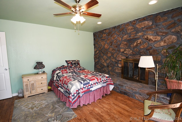 bedroom featuring a stone fireplace, dark hardwood / wood-style flooring, and ceiling fan