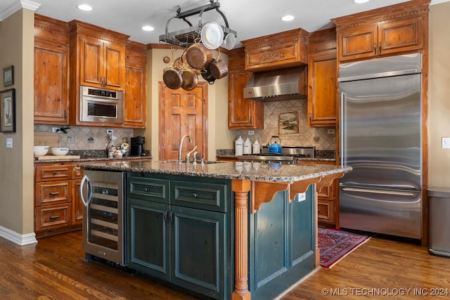 kitchen featuring beverage cooler, a center island with sink, wall chimney range hood, stainless steel appliances, and light stone countertops