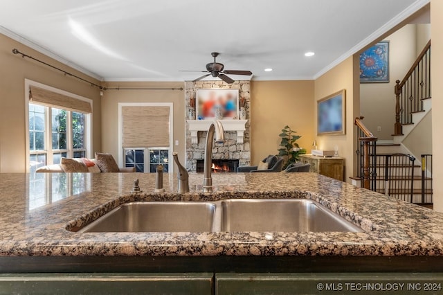 kitchen with ceiling fan, sink, a stone fireplace, dark stone counters, and crown molding