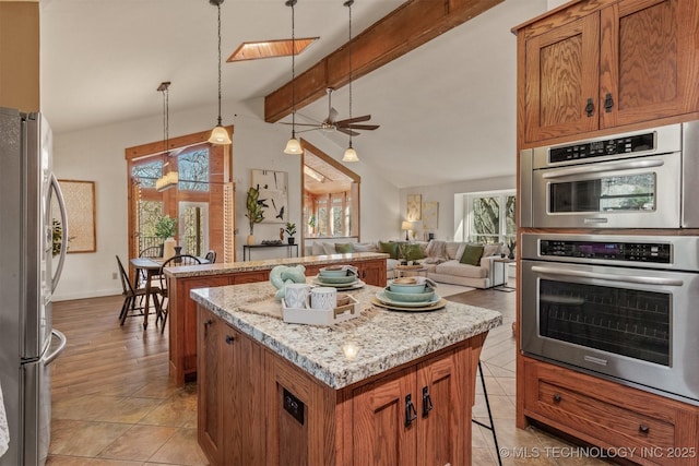 kitchen with lofted ceiling with beams, light stone counters, appliances with stainless steel finishes, a center island, and brown cabinetry