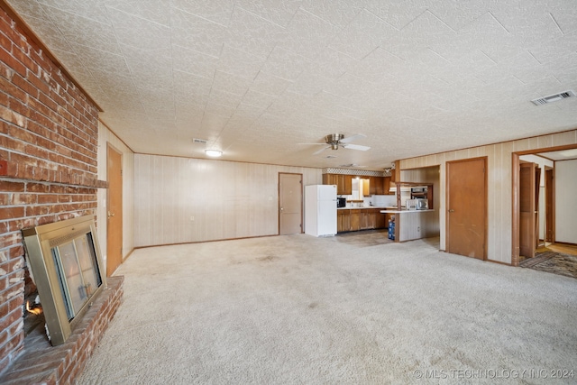 unfurnished living room featuring light carpet, ceiling fan, a textured ceiling, a brick fireplace, and wood walls