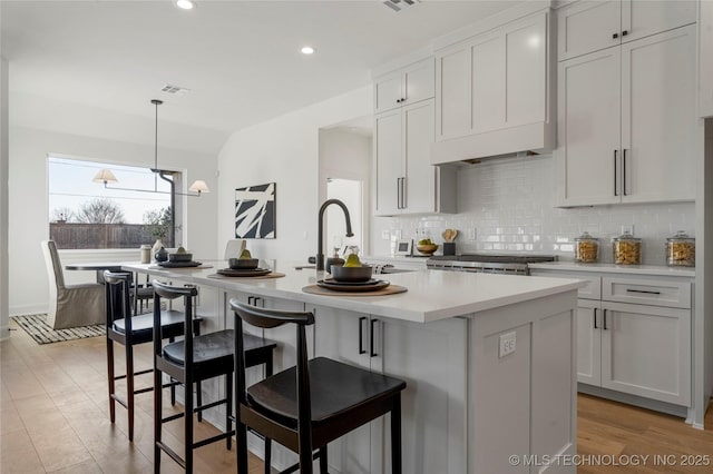 kitchen with decorative backsplash, custom exhaust hood, sink, a center island with sink, and hanging light fixtures