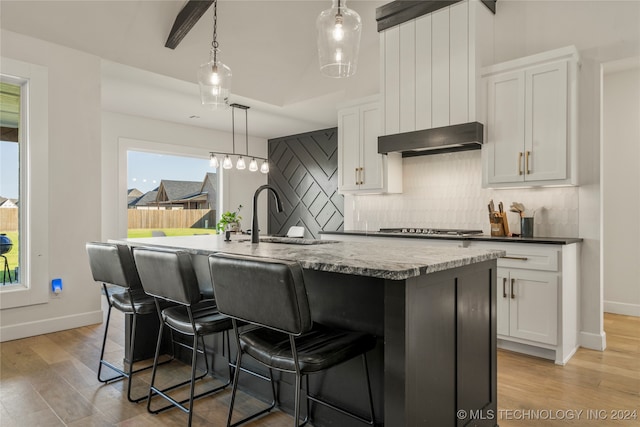 kitchen with light wood-type flooring, a kitchen island with sink, white cabinetry, decorative light fixtures, and backsplash