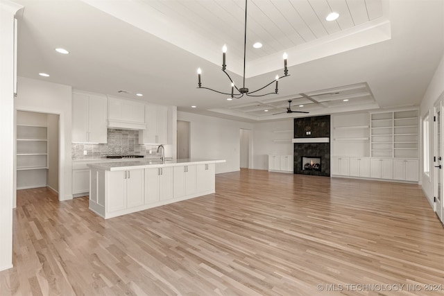 kitchen with an island with sink, a raised ceiling, a tiled fireplace, white cabinetry, and light hardwood / wood-style floors