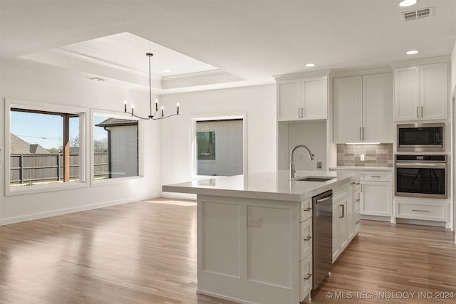 kitchen with a tray ceiling, a center island with sink, light countertops, appliances with stainless steel finishes, and white cabinetry