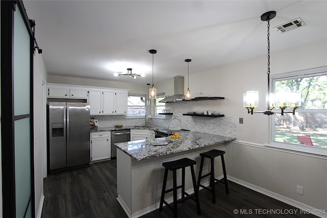 kitchen featuring kitchen peninsula, wall chimney exhaust hood, stainless steel appliances, pendant lighting, and white cabinets