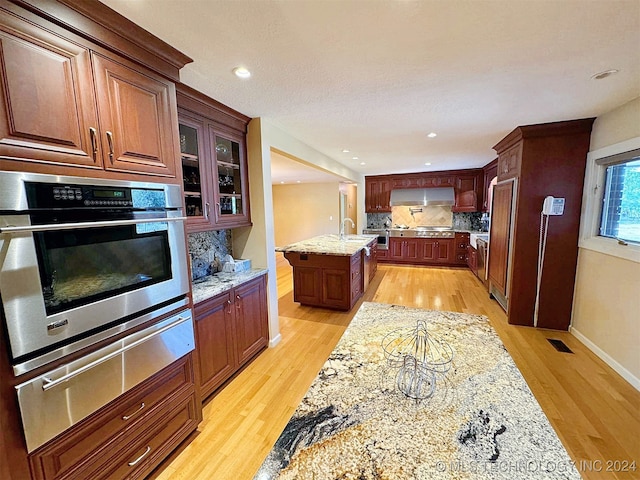 kitchen featuring stainless steel oven, a kitchen island with sink, light hardwood / wood-style flooring, light stone counters, and tasteful backsplash