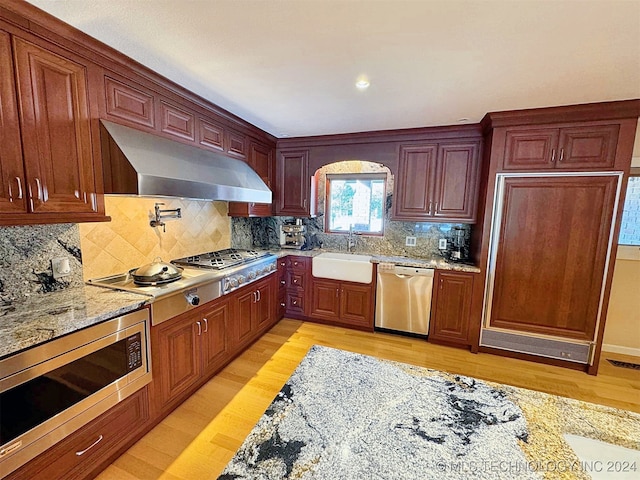 kitchen featuring wall chimney range hood, tasteful backsplash, appliances with stainless steel finishes, light wood-type flooring, and sink