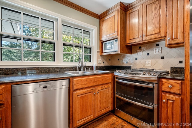 kitchen with appliances with stainless steel finishes, wood-type flooring, ornamental molding, dark stone countertops, and sink