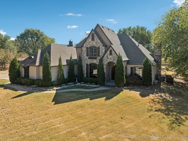 french country home with stucco siding, stone siding, a chimney, and a front lawn