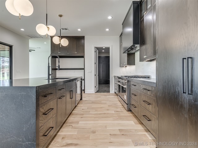 kitchen with wall chimney range hood, an island with sink, stainless steel appliances, sink, and light hardwood / wood-style floors