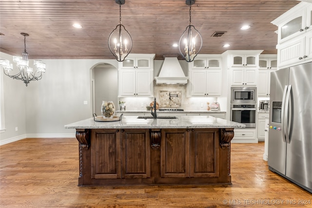 kitchen with an island with sink, stainless steel appliances, decorative light fixtures, custom exhaust hood, and white cabinetry