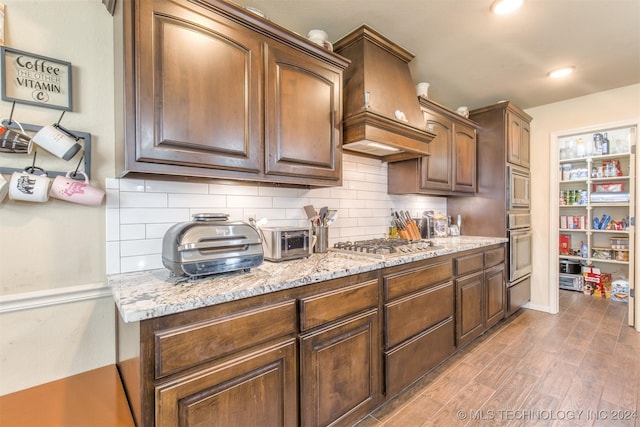 kitchen with light stone countertops, premium range hood, stainless steel appliances, decorative backsplash, and wood finished floors