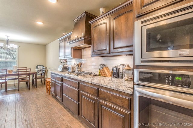 kitchen with appliances with stainless steel finishes, light wood finished floors, a chandelier, backsplash, and custom exhaust hood