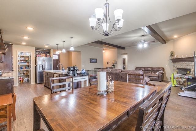 dining room featuring sink, ceiling fan with notable chandelier, a stone fireplace, light hardwood / wood-style floors, and beam ceiling