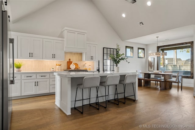 kitchen with an island with sink, hanging light fixtures, wood-type flooring, white cabinets, and high vaulted ceiling