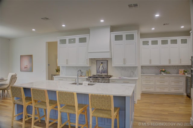 kitchen with tasteful backsplash, light stone counters, a center island with sink, and custom exhaust hood