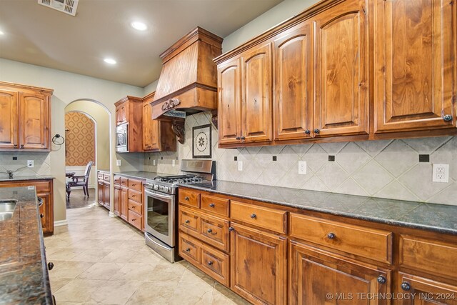 kitchen featuring custom range hood, backsplash, stainless steel appliances, and dark stone countertops