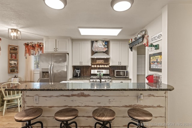 kitchen featuring a kitchen bar, kitchen peninsula, stainless steel appliances, wall chimney exhaust hood, and white cabinets