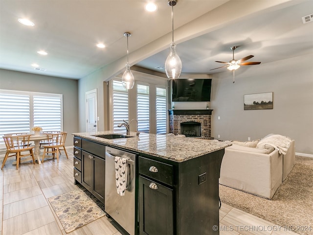 kitchen with a kitchen island with sink, stainless steel dishwasher, light stone countertops, a brick fireplace, and pendant lighting