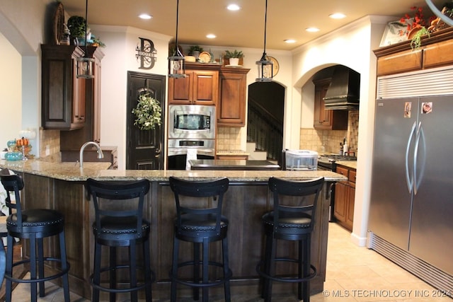 kitchen with tasteful backsplash, light stone counters, built in appliances, a peninsula, and ventilation hood