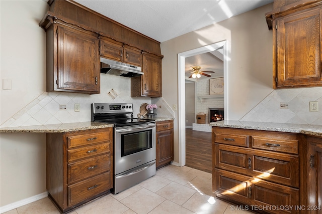 kitchen with ceiling fan, under cabinet range hood, stainless steel electric range, a brick fireplace, and light tile patterned flooring