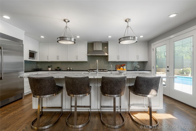 kitchen featuring a center island with sink, built in refrigerator, hanging light fixtures, and wall chimney exhaust hood