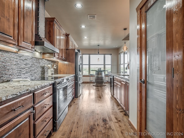 kitchen featuring wall chimney exhaust hood, light hardwood / wood-style flooring, hanging light fixtures, stainless steel appliances, and light stone countertops