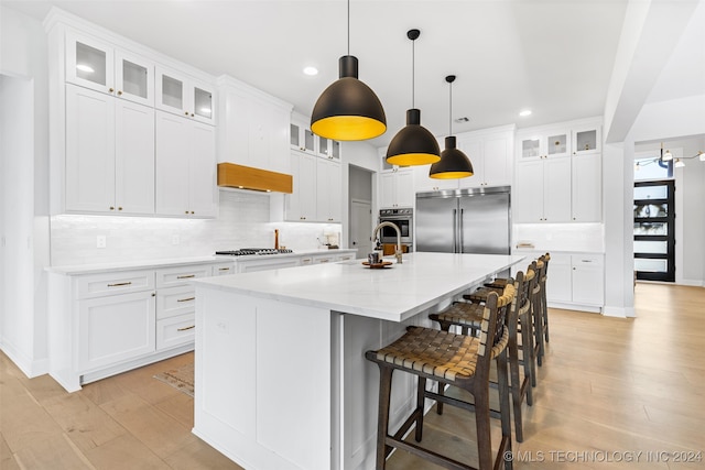 kitchen with appliances with stainless steel finishes, hanging light fixtures, light hardwood / wood-style floors, white cabinets, and a kitchen island with sink