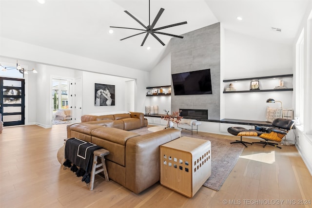 living room featuring ceiling fan with notable chandelier, high vaulted ceiling, a tile fireplace, and light wood-type flooring