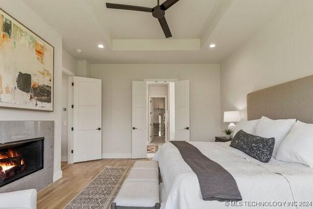 bedroom featuring a raised ceiling, a tiled fireplace, light wood-type flooring, and ceiling fan