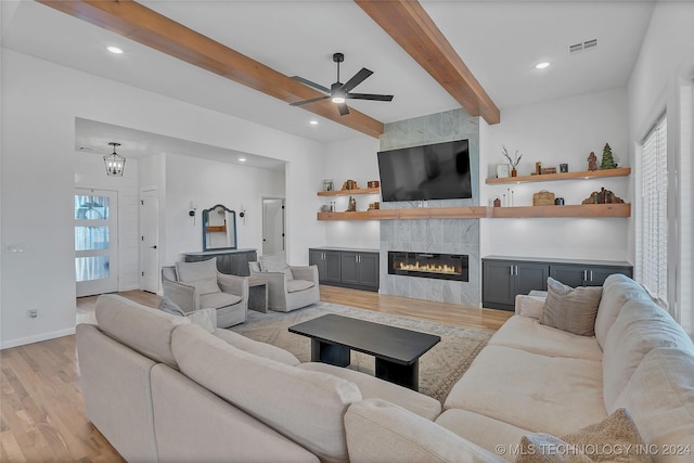 living room with beam ceiling, a tiled fireplace, ceiling fan with notable chandelier, and light wood-type flooring