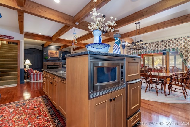 kitchen with beam ceiling, stainless steel microwave, a chandelier, a brick fireplace, and light hardwood / wood-style floors