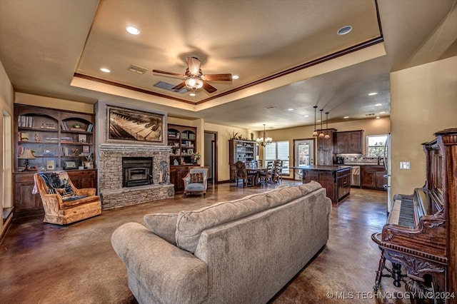 living room featuring a stone fireplace, ceiling fan with notable chandelier, and a raised ceiling