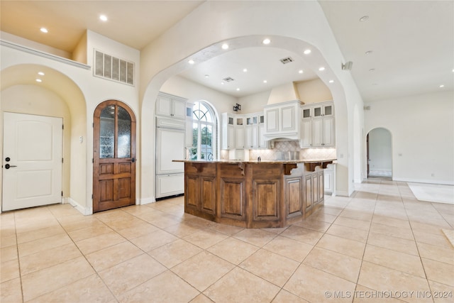 kitchen featuring an island with sink, backsplash, a high ceiling, light stone counters, and light tile patterned floors