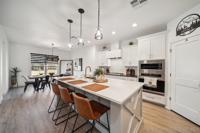 kitchen with appliances with stainless steel finishes, hanging light fixtures, white cabinets, light hardwood / wood-style flooring, and a kitchen island with sink