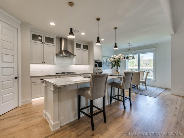 kitchen with light wood-style flooring, a sink, decorative backsplash, wall chimney exhaust hood, and a center island with sink