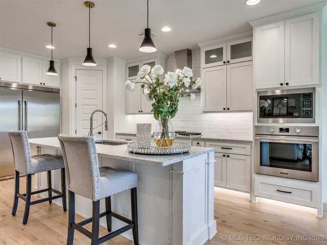 kitchen with built in appliances, light stone counters, white cabinets, and a kitchen island with sink