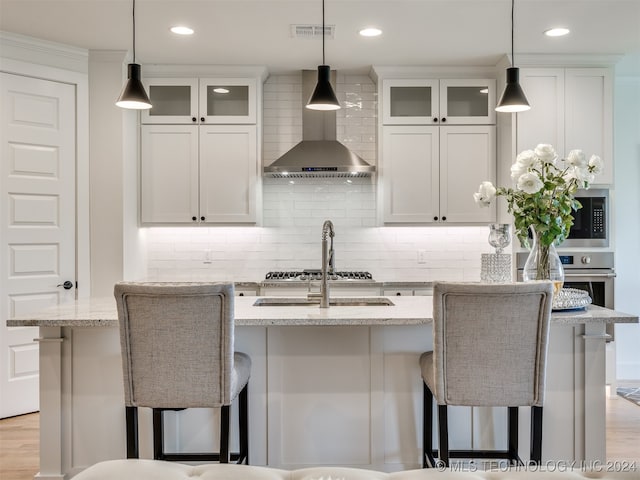 kitchen with wall chimney range hood, white cabinets, light stone countertops, and a center island with sink