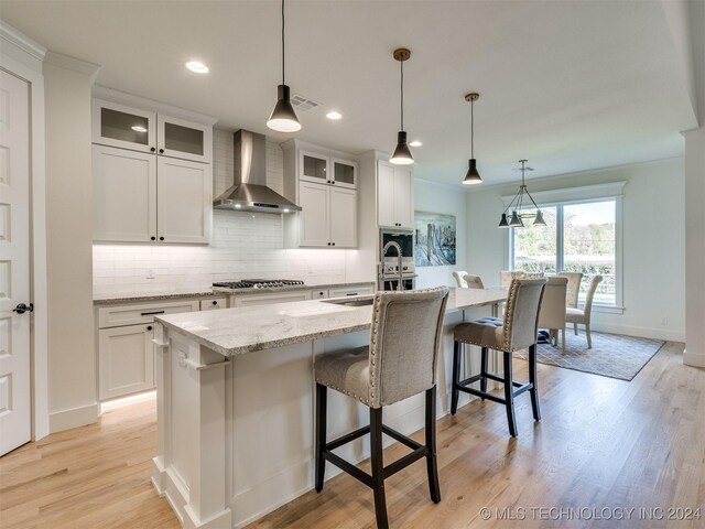 kitchen with wall chimney range hood, a center island with sink, pendant lighting, white cabinets, and light hardwood / wood-style floors