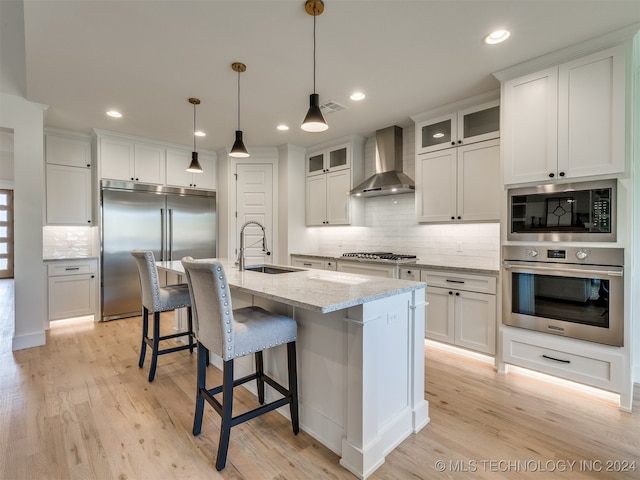 kitchen featuring wall chimney range hood, white cabinetry, built in appliances, light hardwood / wood-style flooring, and a kitchen island with sink